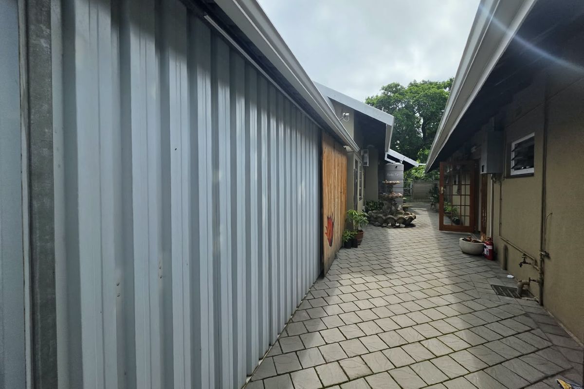 A paved side walkway between a corrugated metal structure and the main house, featuring guttering and an external water tap.
