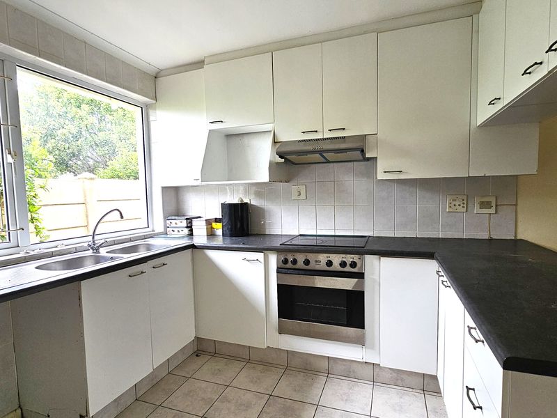 Functional kitchen featuring white cabinetry, dark laminate countertops, built-in oven with hob, extractor fan, and tiled flooring with a large window overlooking the garden.