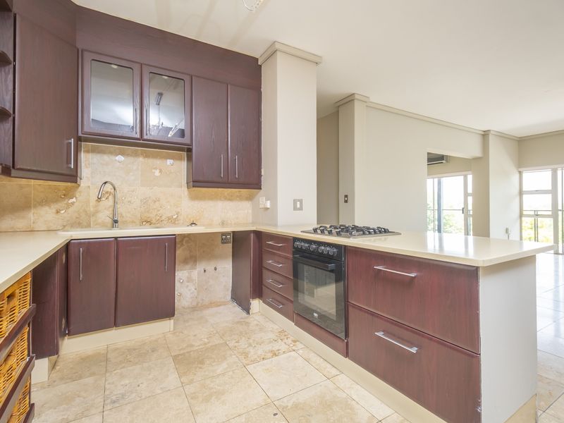 A functional kitchen featuring dark wood cabinetry, stone tile backsplash, built-in oven, gas hob, and stone countertops, opening into a bright living area.