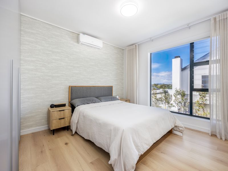 A bright and modern bedroom featuring timber flooring, textured wallpaper, a built-in wardrobe, and an air conditioning unit with large window views.