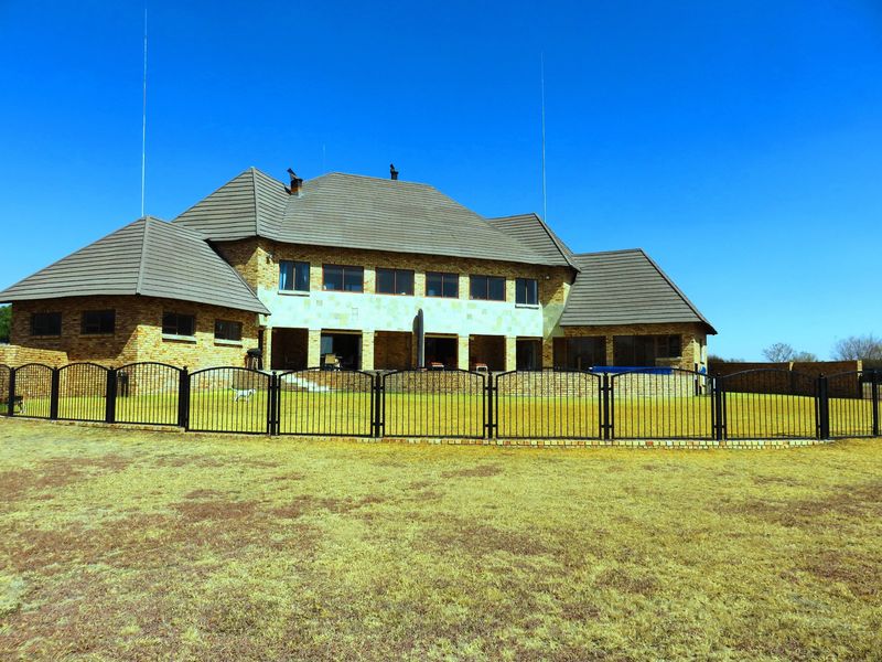 Rear view of a large facebrick small holding residence featuring a tiled roof, covered patio area, and secure metal perimeter fencing.