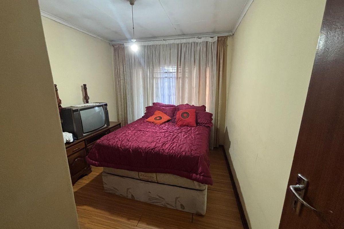 A compact bedroom featuring laminate flooring and large window curtains. The ceiling shows visible signs of wear and staining.