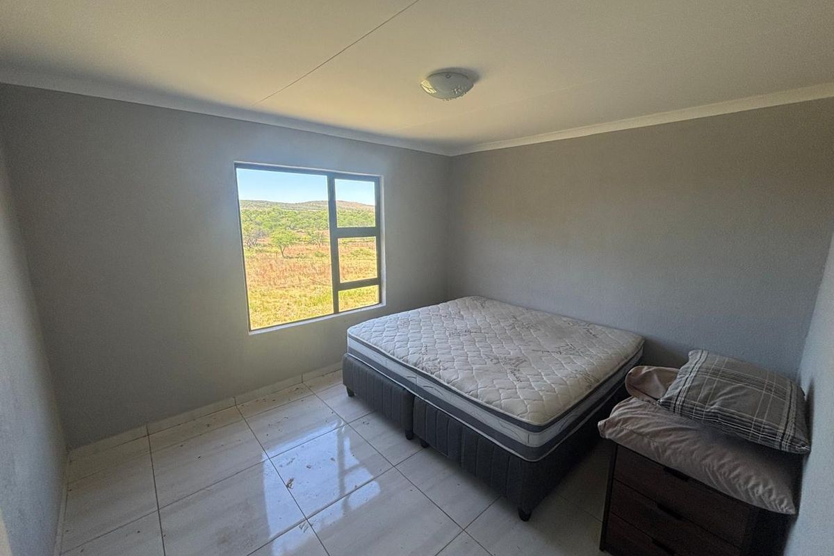 A simple bedroom featuring tiled flooring, neutral grey walls, and a window offering scenic landscape views.