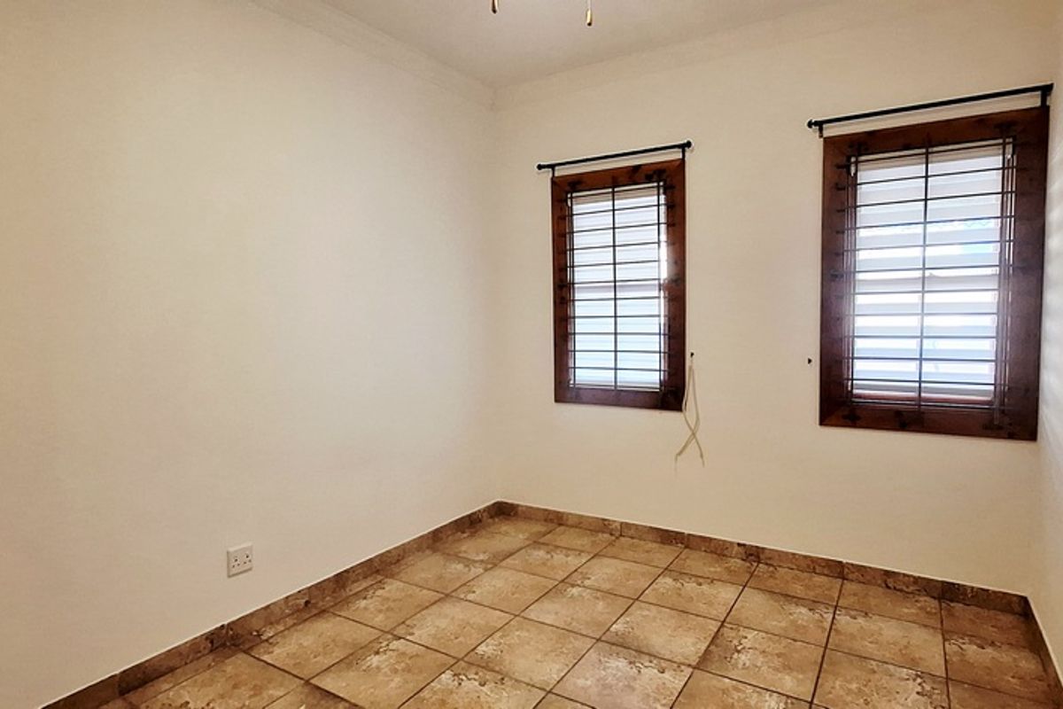 A bright, empty bedroom featuring tiled flooring and two windows with dark wood frames and security bars.
