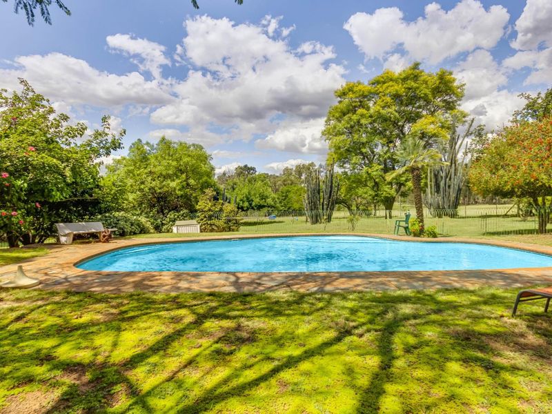 A spacious outdoor pool area set within a lush, green garden landscape, featuring a classic pool design with stone paving.