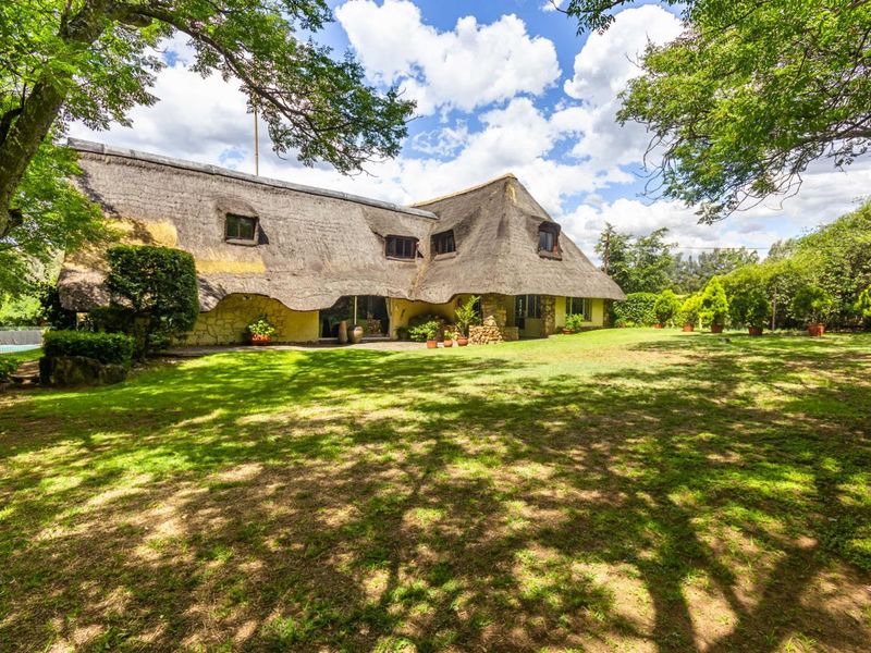 A rear view of a traditional farmhouse featuring a distinctive thatched roof, stone exterior walls, and a spacious, well-maintained garden setting.