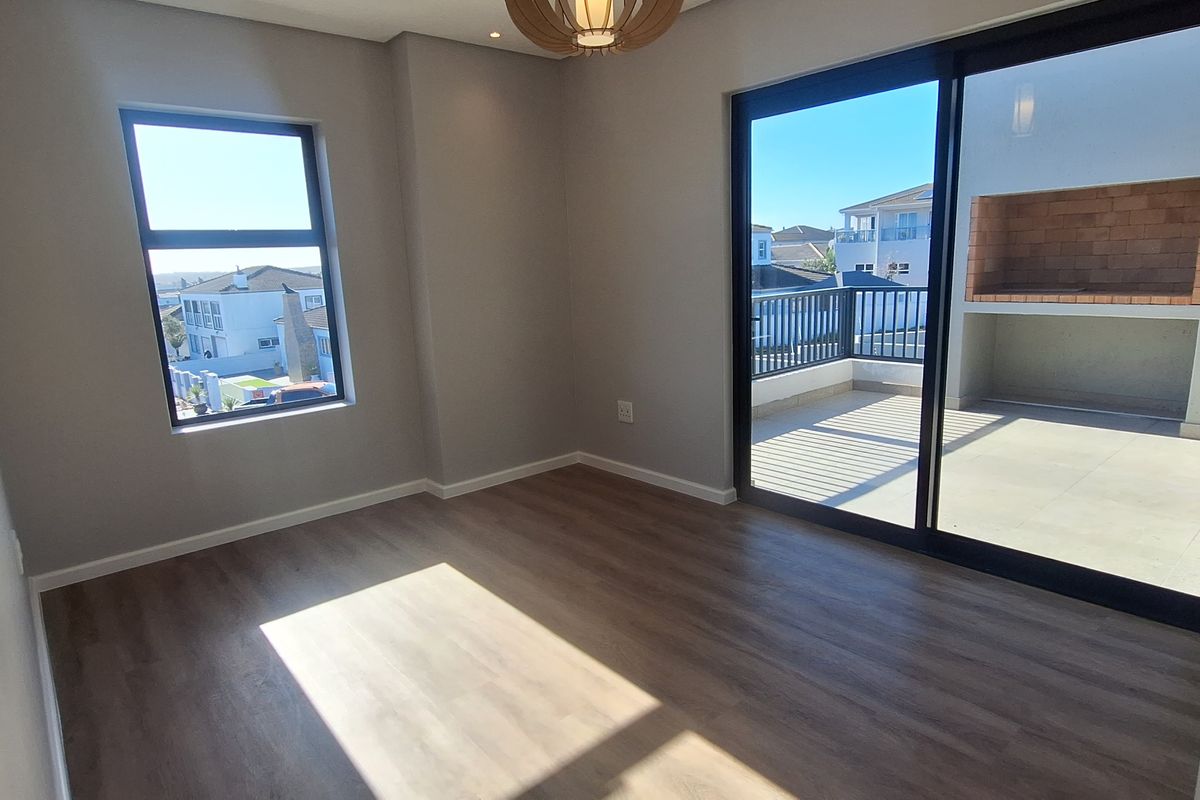 A bright, modern bedroom featuring wood-look flooring, recessed lighting, and large sliding glass doors that open onto a private balcony with a built-in braai.