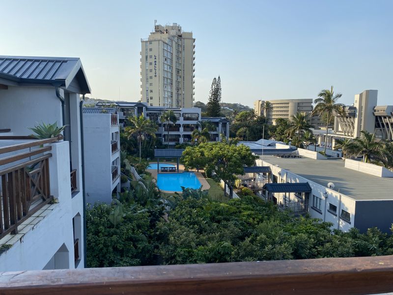 Elevated view overlooking the apartment complex grounds, featuring a swimming pool, lush garden areas, and surrounding residential buildings.