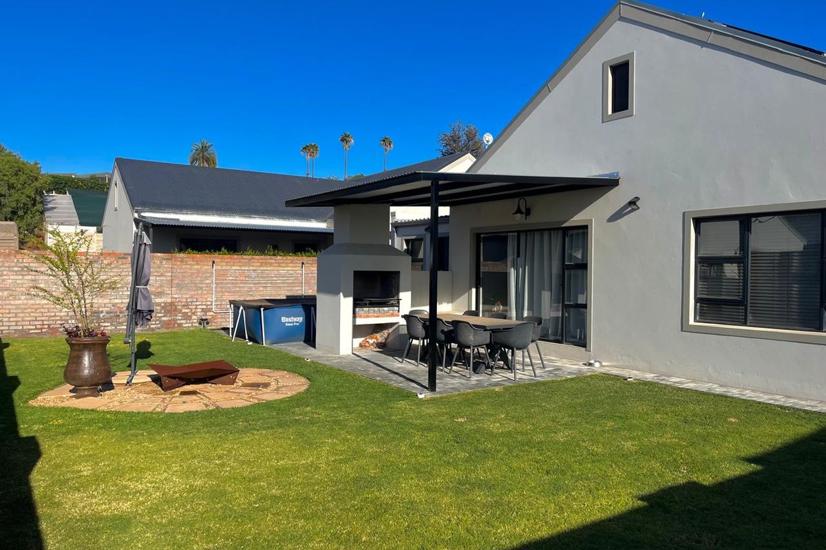 A well-maintained rear exterior featuring a covered patio with a built-in braai, paved flooring, and a neat garden area with brick boundary walls.