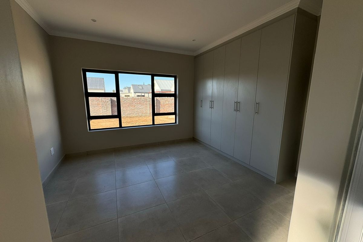 A clean, modern bedroom featuring neutral tiled flooring, built-in cupboards, and large aluminium-framed windows providing natural light.