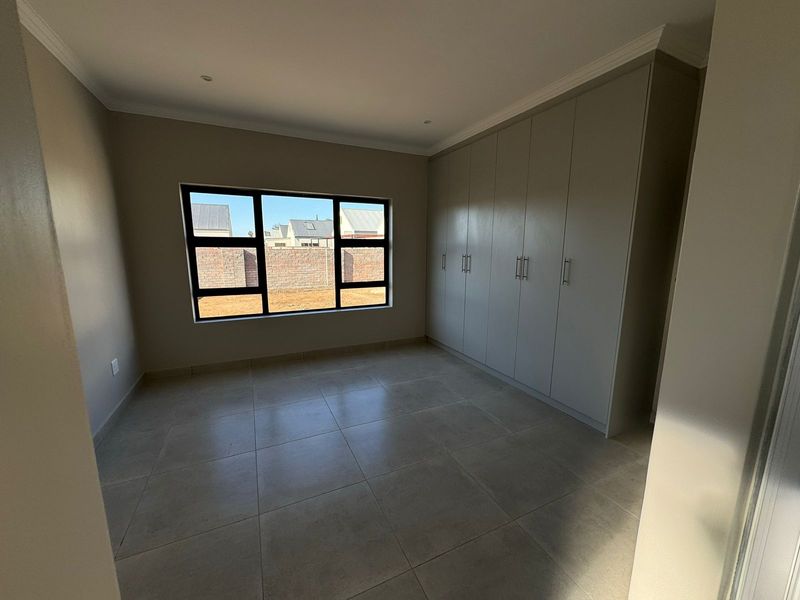 A clean, modern bedroom featuring neutral tiled flooring, built-in cupboards, and large aluminium-framed windows providing natural light.