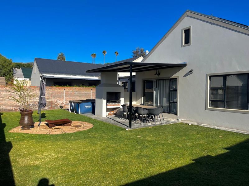 A well-maintained rear garden featuring a covered patio with a built-in braai, paved entertainment area, and established lawn.