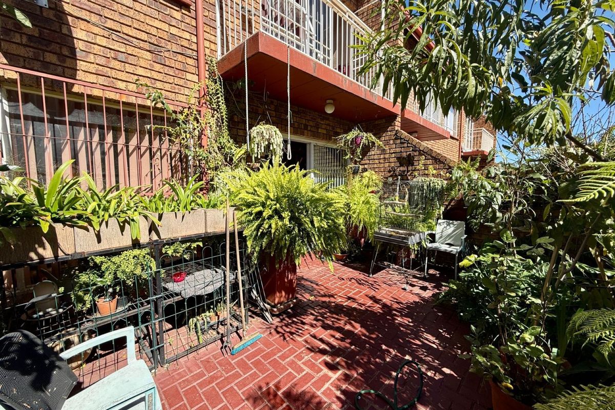 A private rear patio area featuring red brick paving, established garden greenery, and an overhead balcony with metal security railings.