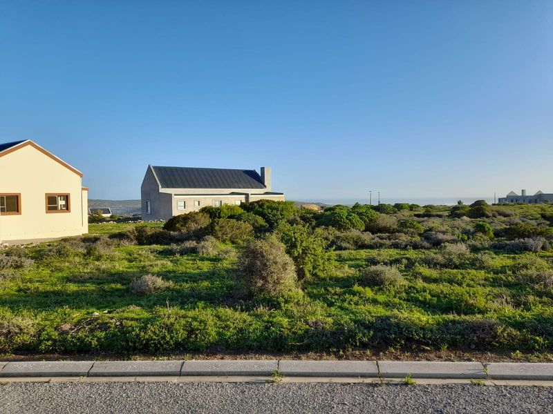 A view of a vacant residential plot featuring native coastal vegetation, adjacent to a paved road with established neighboring properties in the background.
