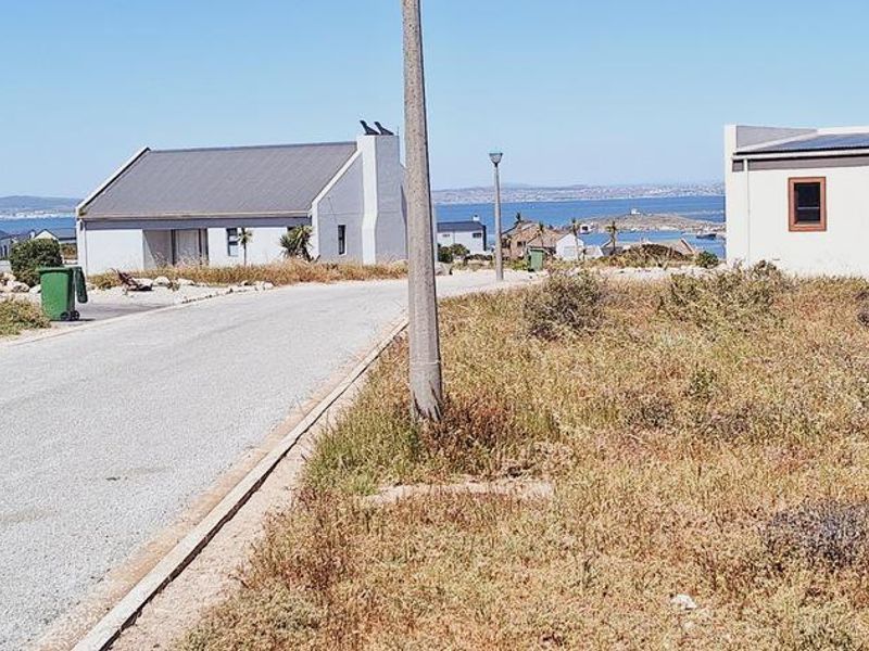 A street view of a residential area featuring a paved road, utility infrastructure, and surrounding coastal homes with distant ocean views.