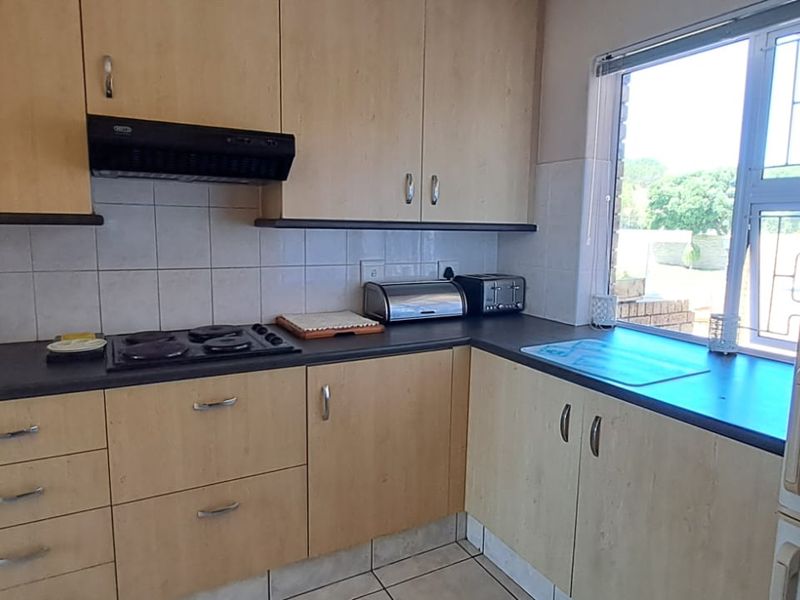 A functional kitchen featuring light wood-finish cabinetry, dark countertops, an electric hob with extractor fan, and tiled flooring with a window view.