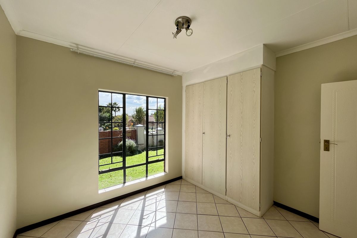 A bright, empty bedroom featuring neutral tiled flooring, built-in wooden cupboards, and a large window overlooking the garden.