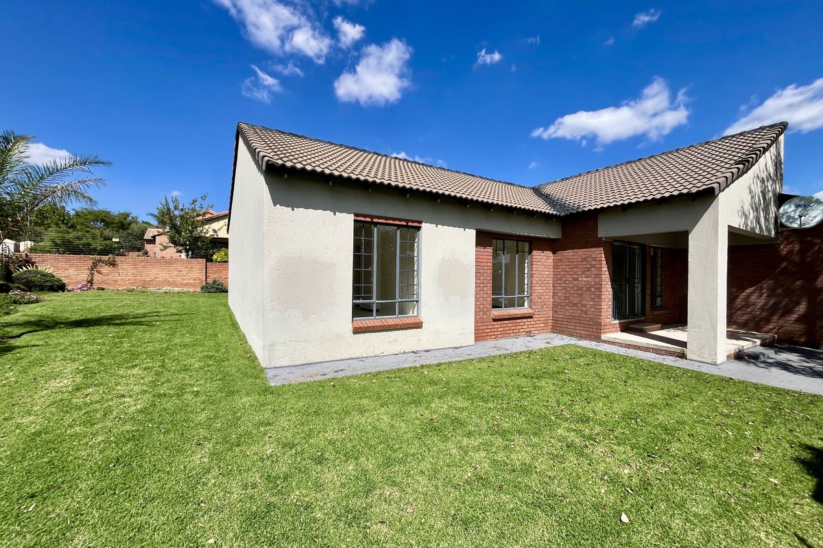 Rear view of a single-story house featuring a brick and plastered facade, a covered patio area, and a well-maintained lawn with brick boundary walls.