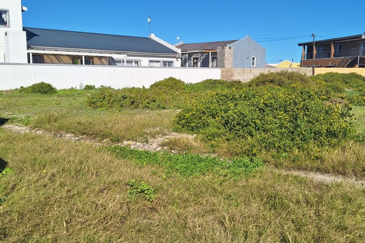 A vacant residential plot featuring natural coastal vegetation, enclosed by a perimeter boundary wall with established neighboring properties in the background.