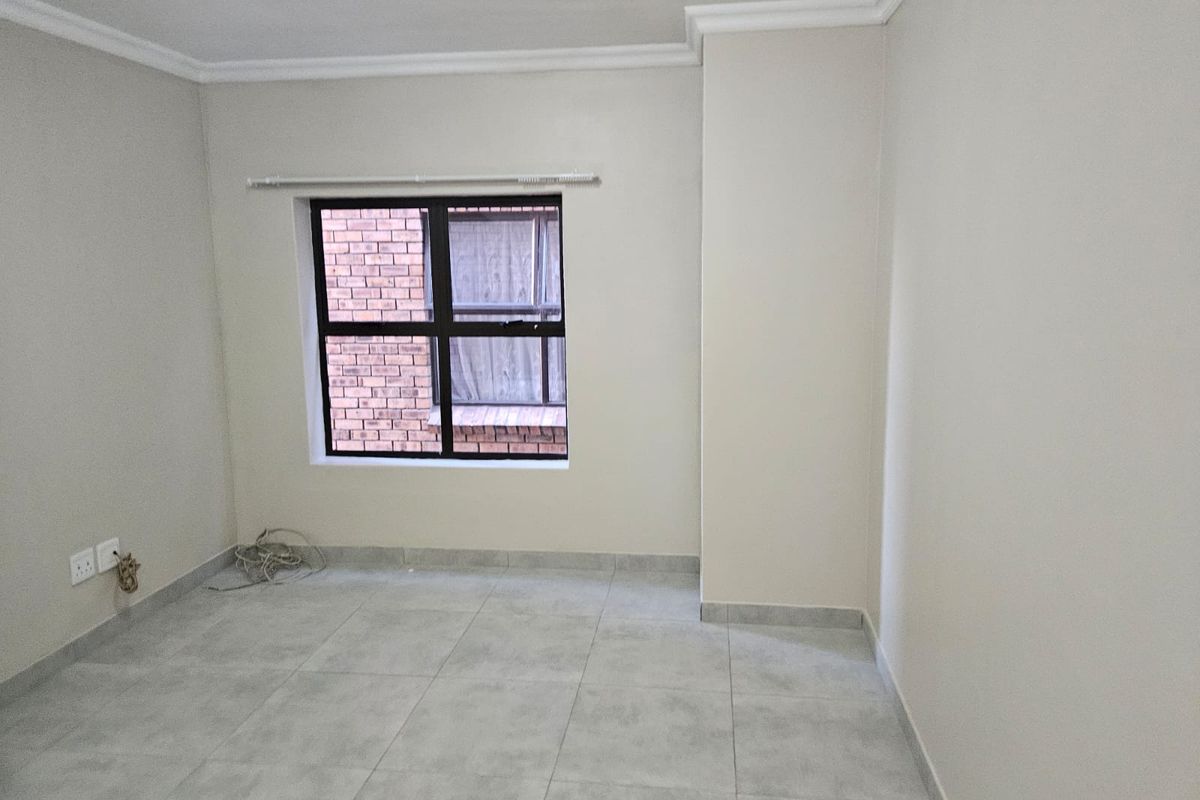 A clean, empty bedroom featuring neutral tiled flooring, white walls with crown molding, and a dark aluminium window frame.