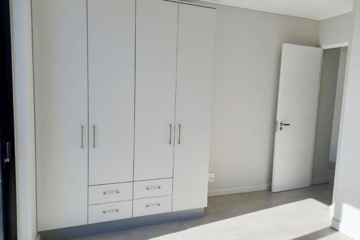 A bright, empty bedroom featuring neutral tiled flooring and a modern, white built-in wardrobe with integrated drawers.