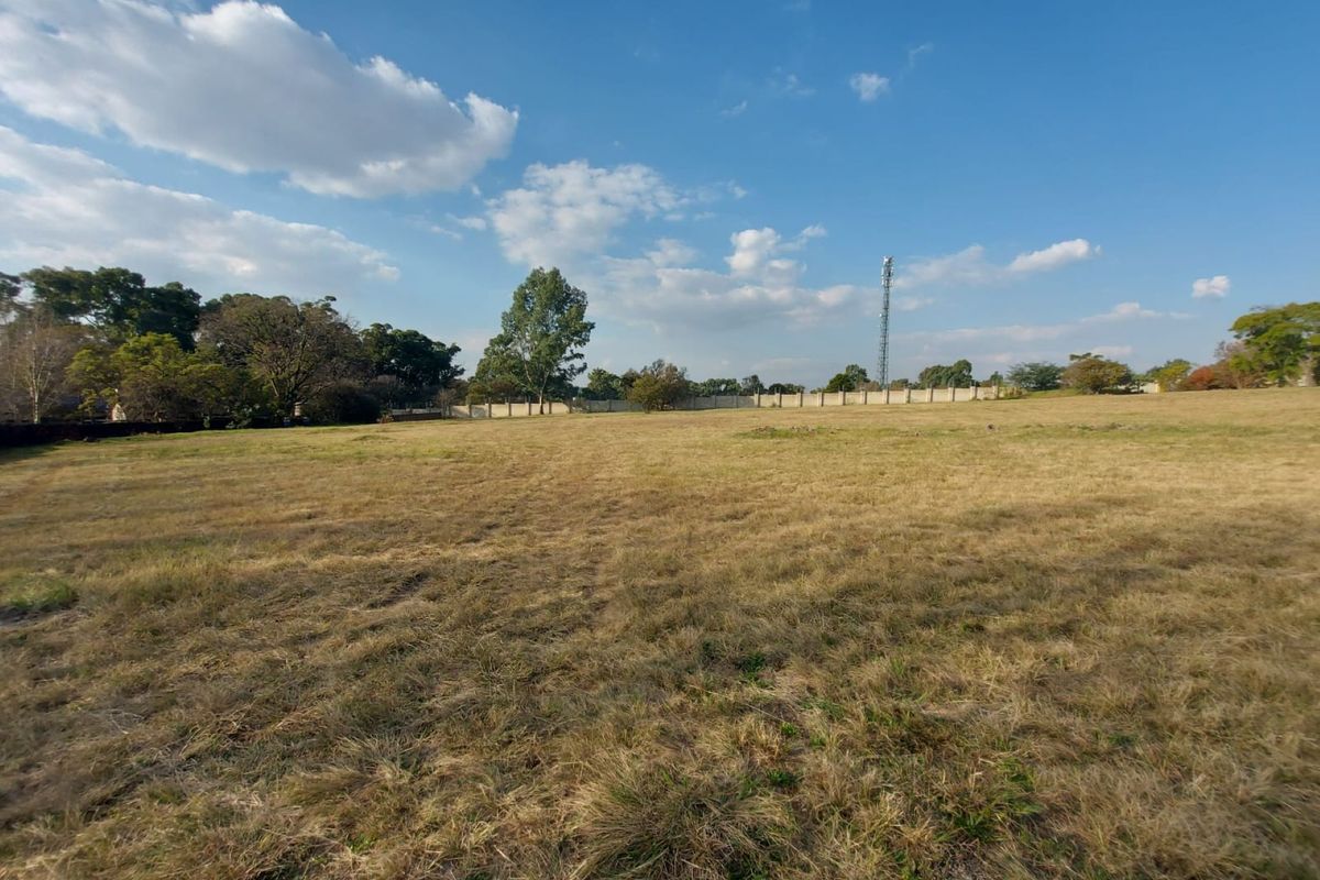 A wide-angle view of a vacant residential plot featuring dry grass, perimeter boundary walls, and mature trees in the background.