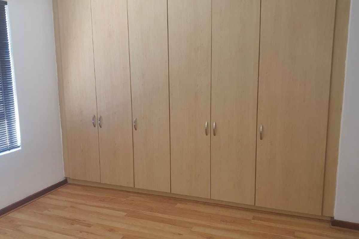 A clean bedroom featuring light wood-finish built-in cupboards, laminate flooring, and a window fitted with venetian blinds.