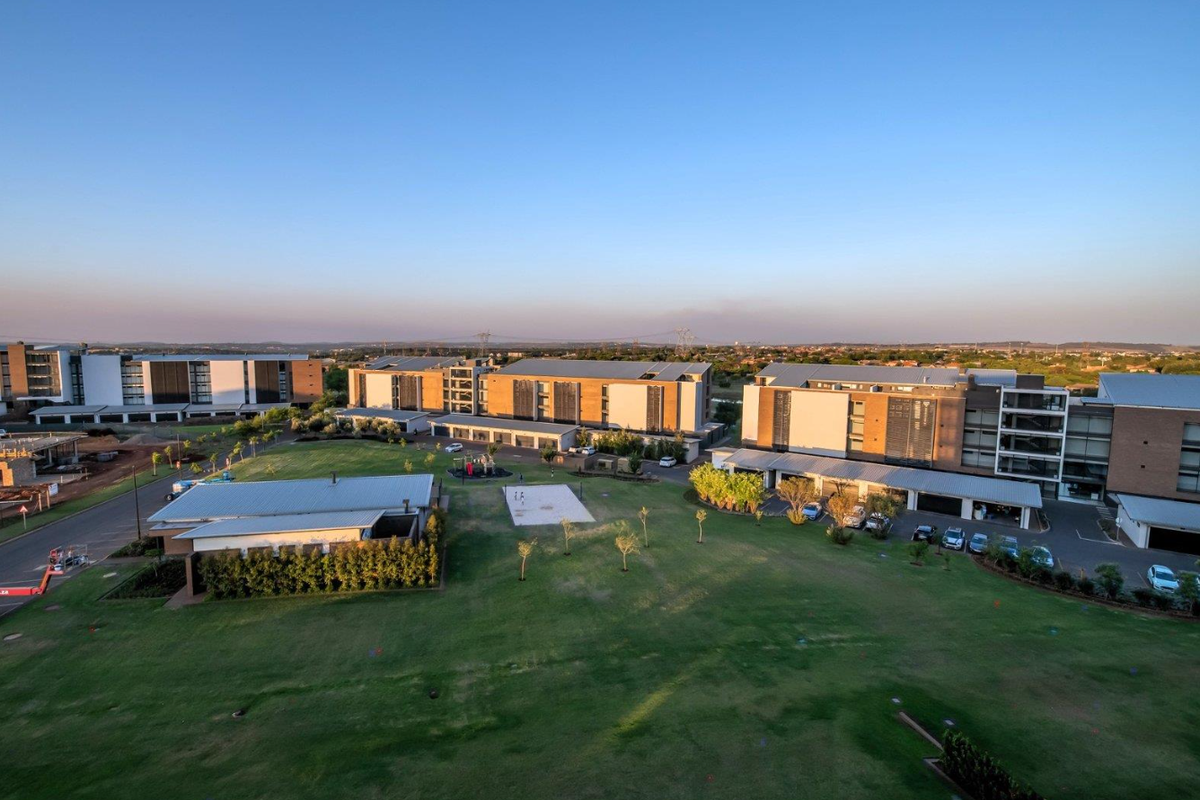 An elevated aerial view of a modern apartment complex featuring landscaped green spaces, paved parking areas, and contemporary multi-story residential buildings.