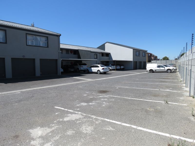 Exterior view of an apartment complex featuring a paved parking area, covered carports, and secure boundary walling with electric fencing.