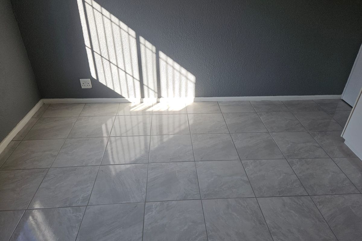 A bright, empty bedroom featuring neutral grey textured walls and modern light-toned tiled flooring with natural light streaming in.