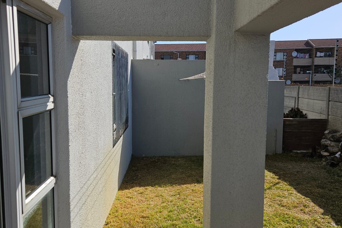 A rear exterior view of the apartment featuring a small lawn, paved patio area, and a structural concrete pergola.