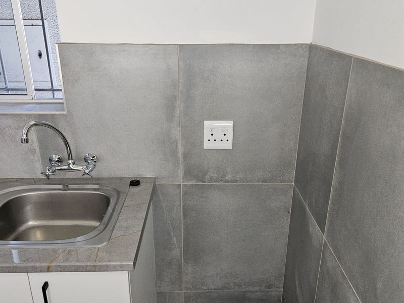 A clean, modern laundry area featuring a stainless steel sink, chrome mixer tap, stone-look countertop, and contemporary grey wall tiling.