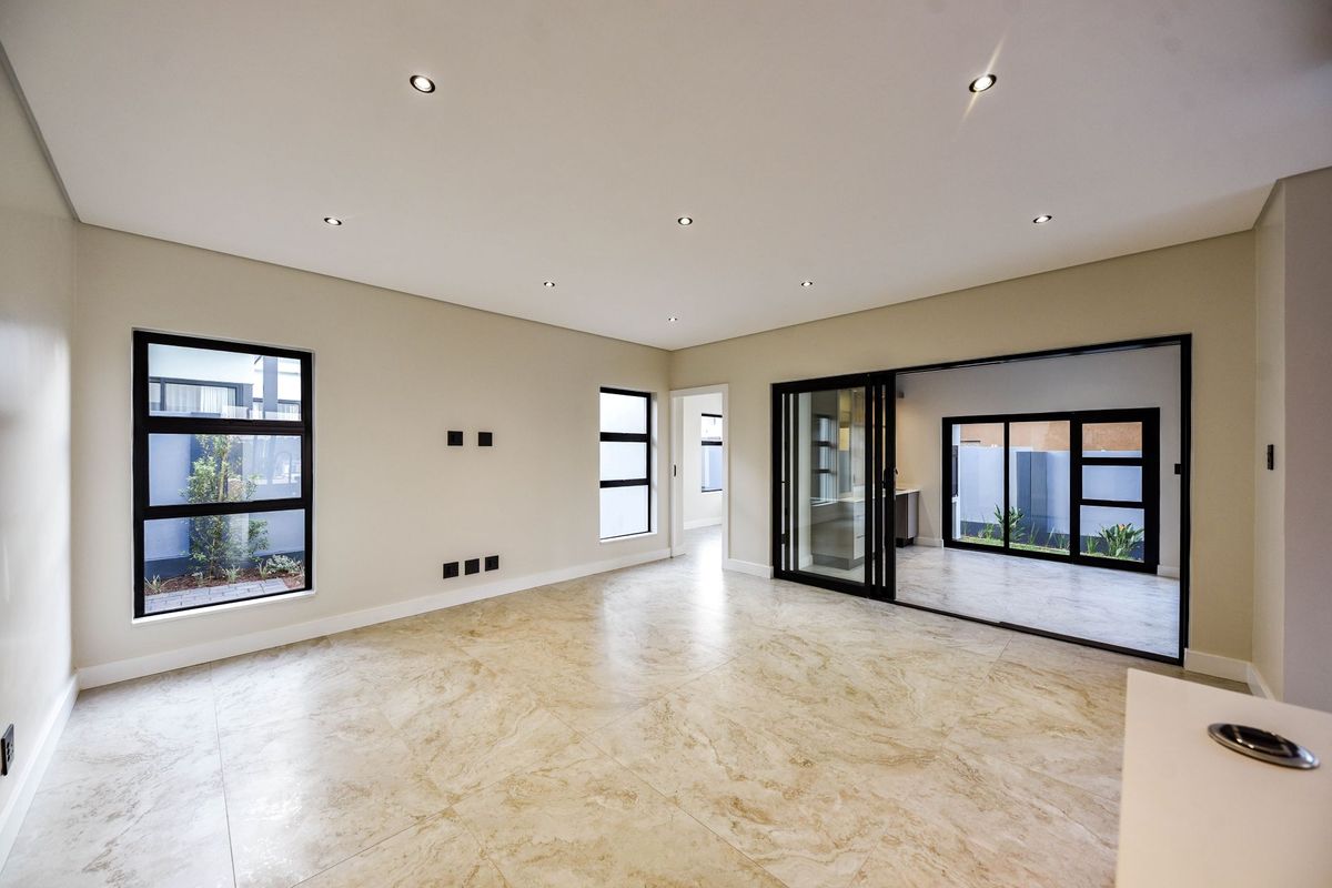 A bright, modern living area featuring large format floor tiles, recessed downlighting, and black-framed sliding doors opening to an outdoor patio space.