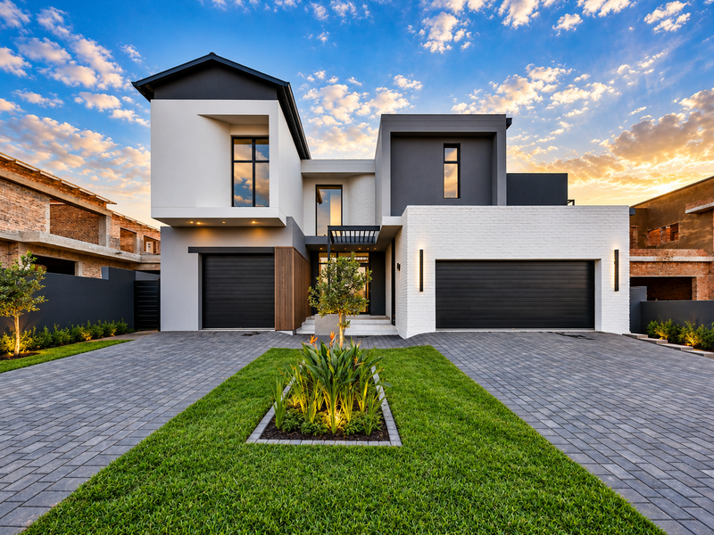 A stunning modern double-story home featuring a paved driveway, double garage, and beautifully landscaped front garden under a vibrant sunset sky.
