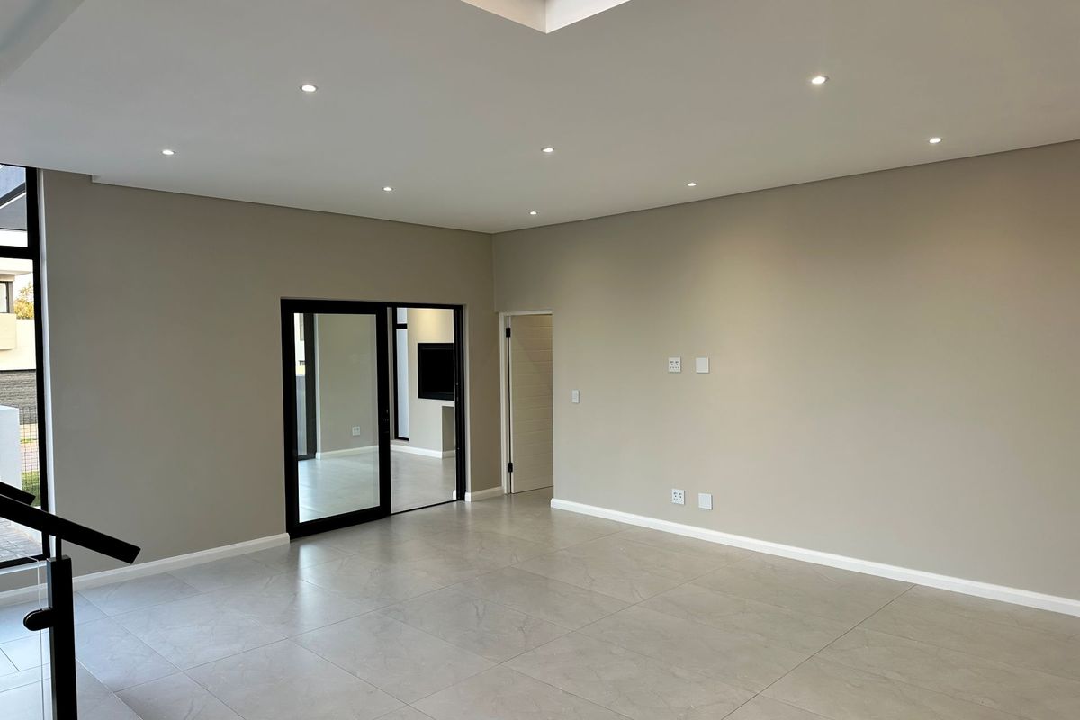 A spacious, modern living area featuring neutral tones, large format floor tiles, recessed ceiling lighting, and a sleek staircase with a glass balustrade.