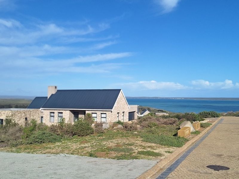 A coastal house featuring stone cladding and a dark metal roof, situated in a scenic landscape with ocean views.