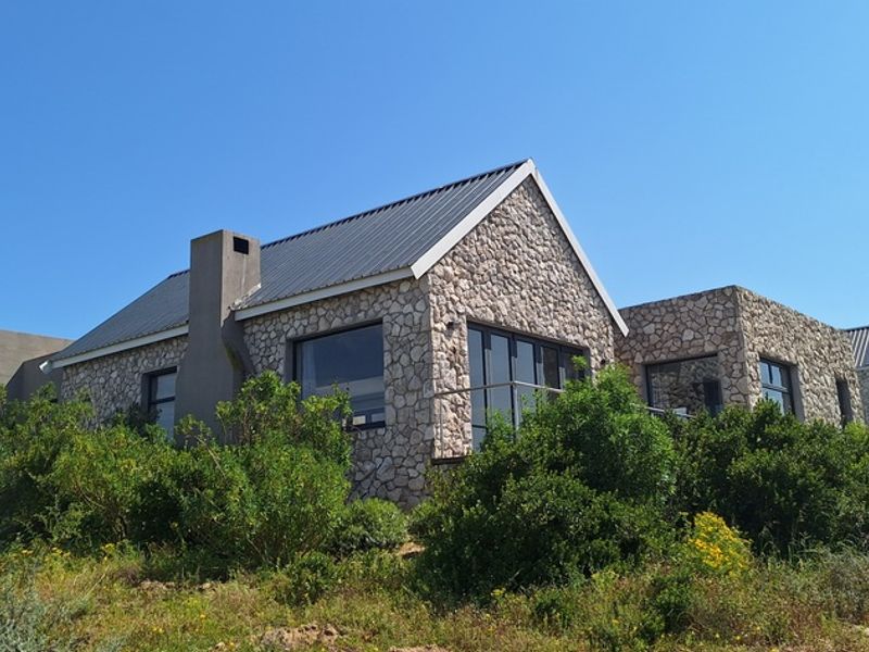A rear view of a modern house featuring stone cladding, a metal roof, and an external chimney, set within a landscaped coastal environment.