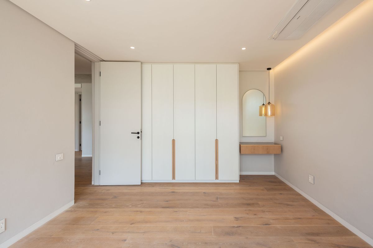 A modern bedroom featuring light wood flooring, built-in floor-to-ceiling wardrobes, a floating vanity shelf, and integrated ceiling lighting.