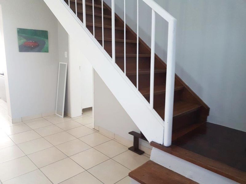 A bright hallway featuring a wooden staircase with a white balustrade and neutral ceramic floor tiles.