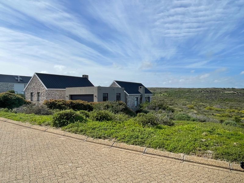 A view of a modern stone-clad residential property with a dark metal roof, situated in a natural coastal landscape setting.