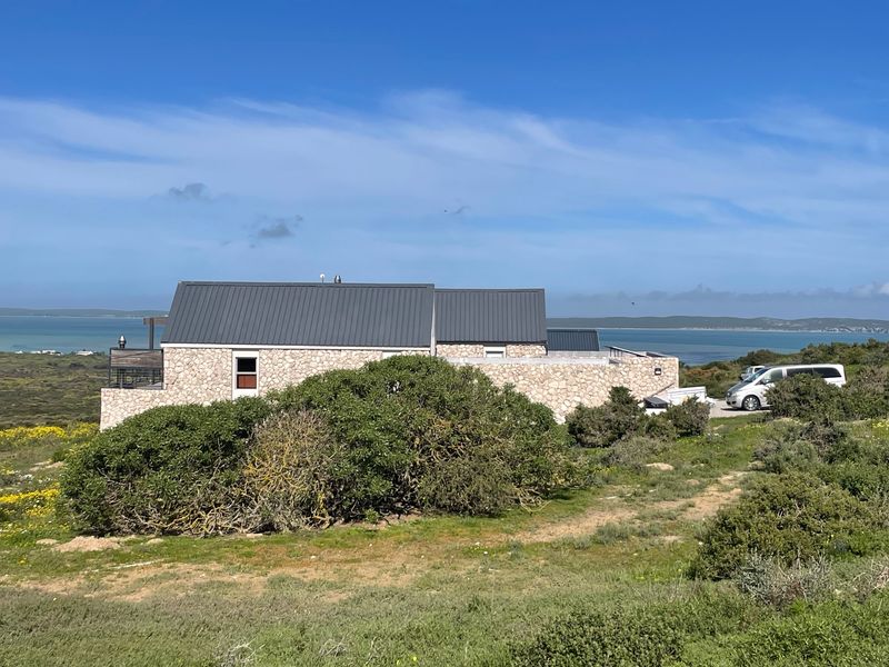A view of the rear elevation of a stone-clad residence featuring a metal roof, set within a natural coastal landscape with water views.