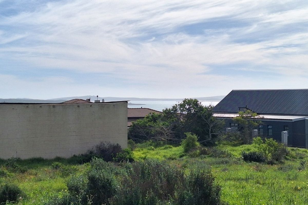 A view of a vacant residential plot featuring natural vegetation, with surrounding residential properties and a distant water view in the background.