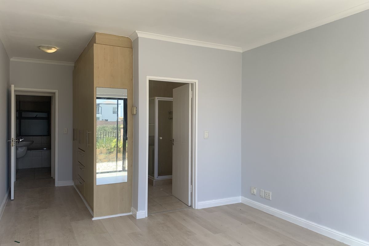 A bright, empty bedroom featuring light-toned laminate flooring, built-in wooden cupboards with a mirror, and direct access to an en-suite bathroom.