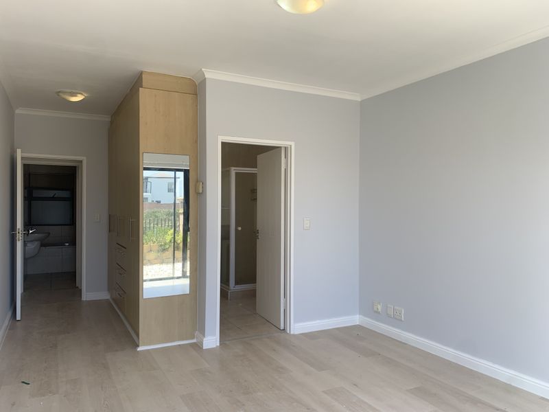 A bright, empty bedroom featuring light-toned laminate flooring, built-in wooden cupboards with a mirror, and direct access to an en-suite bathroom.