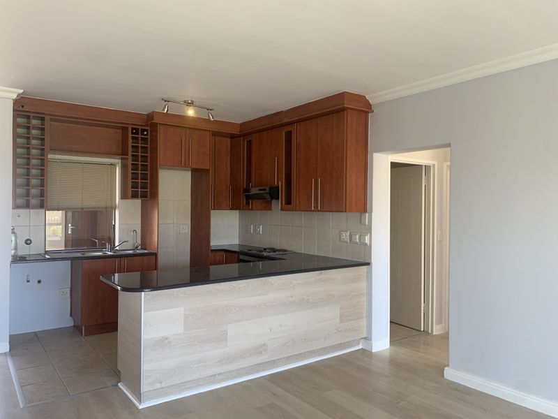 Open plan kitchen featuring wooden cabinetry, granite countertops, a breakfast bar, and integrated hob, seamlessly flowing into the living area.