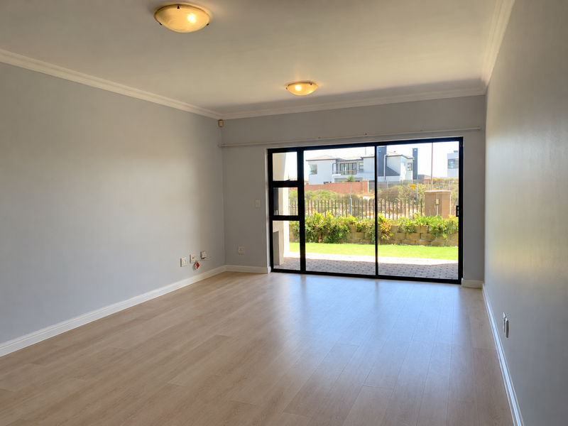 A bright and spacious empty living room featuring modern laminate flooring, neutral walls, and a large sliding glass door providing access to the garden.