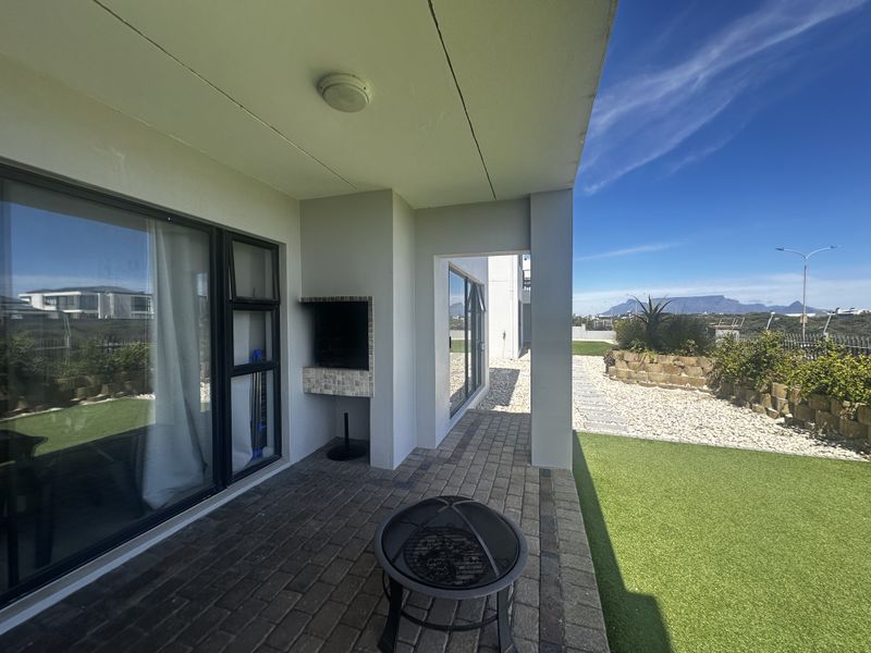 A covered patio area featuring paved flooring, a built-in braai, and sliding glass doors, overlooking a landscaped garden with mountain views.