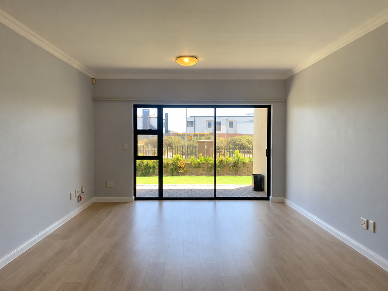 A bright, empty living room featuring light-toned laminate flooring, neutral walls, and a large sliding glass door providing access to the rear garden area.