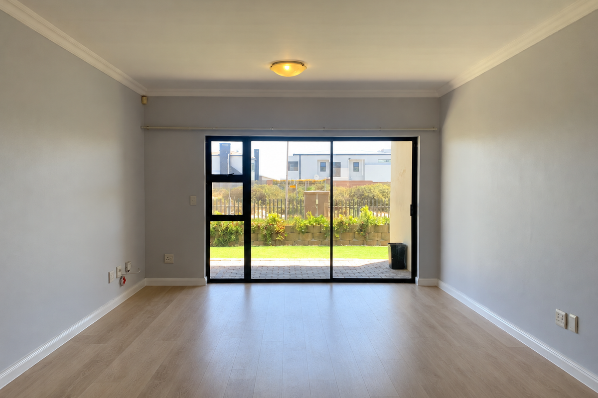 A bright, empty living room featuring light-toned laminate flooring, neutral walls, and a large sliding glass door providing access to the rear garden area.