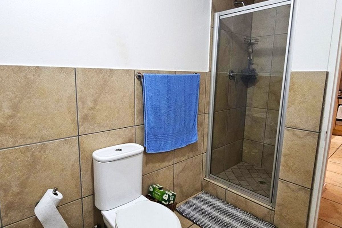 A functional bathroom featuring a glass-enclosed shower, ceramic wall tiling, and a standard toilet, with an adjacent view into the kitchen area.
